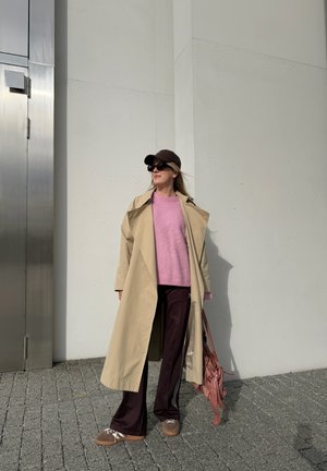 Gabardina beige, suéter rosa, pantalones deportivos oscuros con franjas blancas, gorra marrón, gafas de sol oscuras y zapatos marrones. Bolsa rosa al hombro.
