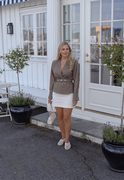 Blusa marrón con detalles de botones, mini falda blanca, zapatos de tela de esparto beige, llevando un pequeño bolso beige, de pie al aire libre.