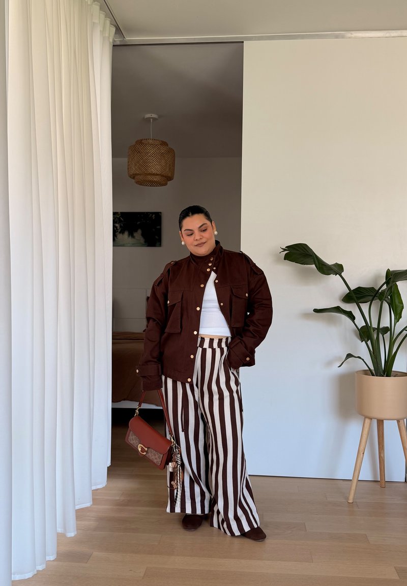 Femme debout à l'intérieur, portant une veste marron et un haut blanc avec un pantalon large rayé marron et blanc, tenant un petit sac à main marron.