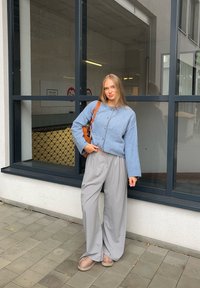 Világoskék kötött pulóver gombos részletekkel, laza szürke nadrággal és barna lábbelivel párosítva, egy nagy üvegablak háttér előtt.