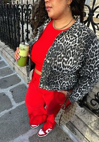 Leopard-print denim jacket, red crop top, red joggers, and red/white trainers. Holding a green drink in a clear cup with a straw.