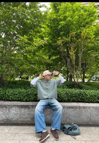 Överdimensionerad skjorta i grön och vit rutigt mönster, ljusblå denimjeans, bruna skor, beige keps, sittande på en stenplatta med grönskande bakgrund.