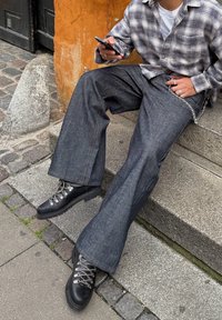 Wijdvallende donkerblauwe spijkerbroek met zichtbare steken, gecombineerd met zwarte wandelschoenen met metalen oogjes en grijze veters, zittend op stenen traptreden.