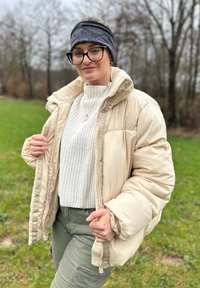 Casaco de penas bege com colarinho alto, combinado com um suéter de malha branco e calças verde-azeitona. Uma faixa cinza na cabeça e um colar completam o visual.