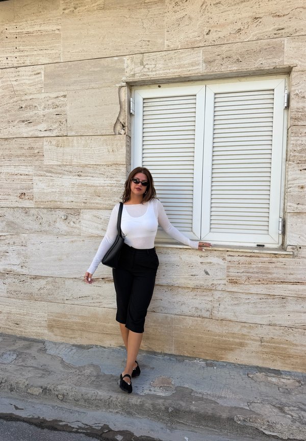 Mujer con gafas de sol, top blanco de manga larga, pantalones negros y zapatos negros, de pie contra una pared de piedra beige con persianas blancas cerradas.