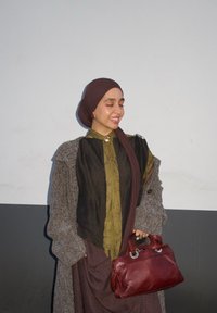 Manteau en laine brun porté sur un chemisier vert olive, jupe brune et hijab assorti. Tenant un sac  à main en cuir rouge avec des accents métalliques.