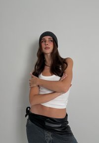 Débardeur blanc, jupe en similicuir noir, jean en denim. Cheveux bruns longs et ondulés, bonnet en maille gris, bras croisés, fond neutre.