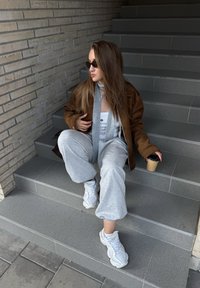Bruine jas, lichtgrijze sweatpants, witte sneakers, grijze sjaal, met een koffiekop in de hand, zittend op grijze stenen treden, bakstenen muur op de achtergrond.