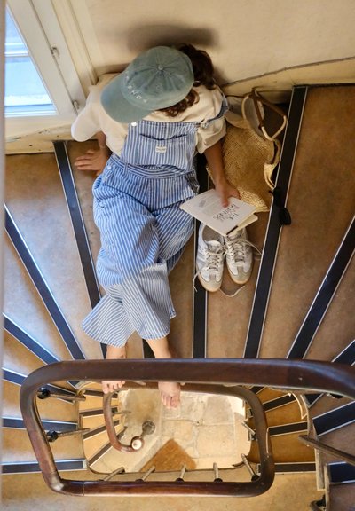 Overoles a rayas azules y camisa blanca, sentado en una escalera con barandilla de madera. Los artículos cercanos incluyen un cuaderno y zapatillas. Luz natural.