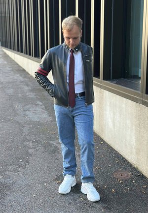 Chaqueta de cuero negra con rayas rojas, camisa blanca, corbata roja, jeans de color azul claro y zapatillas blancas. El fondo presenta paneles de vidrio oscuros.