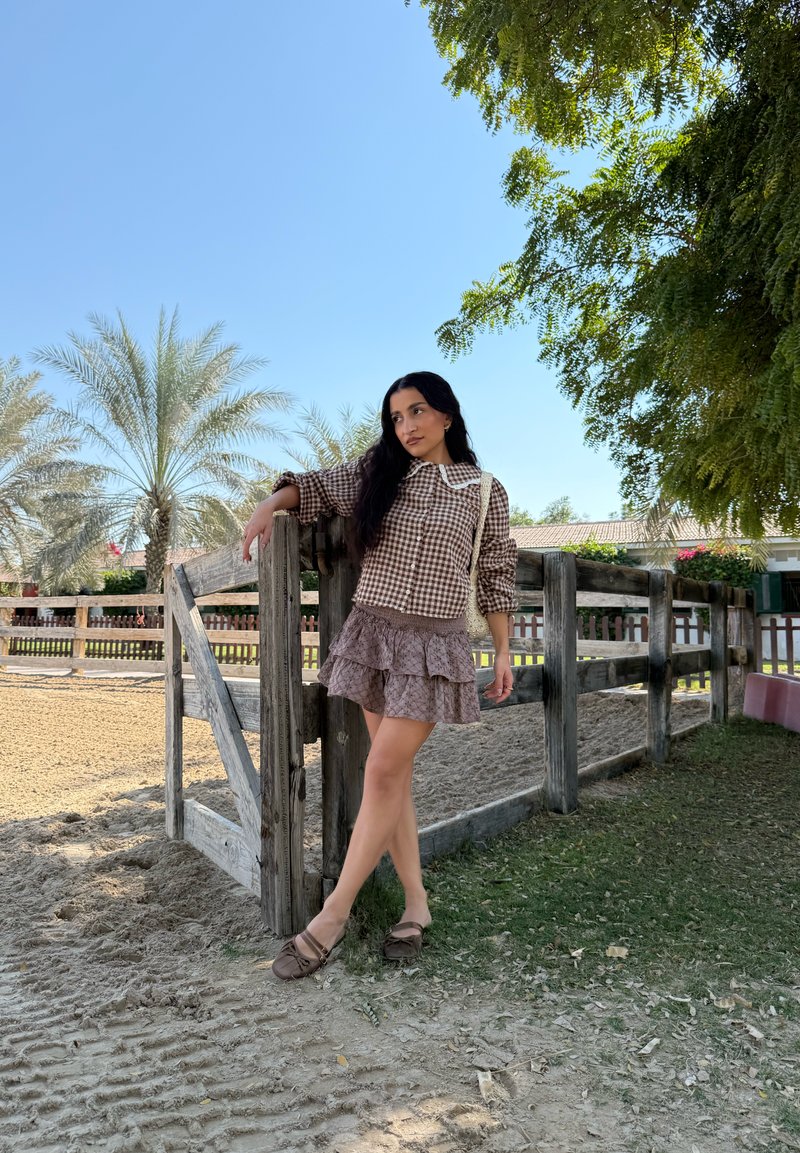 Vrouw in een bruin geruit overhemd en een rok met ruches leunt tegen een houten hek onder een blauwe lucht met palmbomen en groen in de buurt.