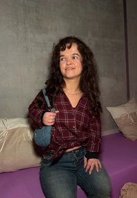 Jeune femme aux cheveux bouclés portant une chemise à carreaux bordeaux et un jean, assise sur un banc violet, tenant un petit sac à main bleu.