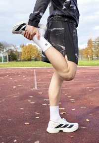 Idrottare i svarta shorts med abstrakt mönster, vita strumpor och vita sneakers med svarta ränder, som stretchar benet på röd löparbana.