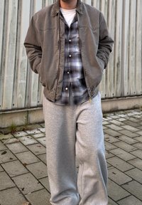 Ruskea pommikoni takki elastisilla hihoilla, kerroksittain sinisen ruudullisen flanellipaidan päällä, yhdistettynä vaaleanharmaisiin verkkareihin. Rakenne näkyy kankaissa.