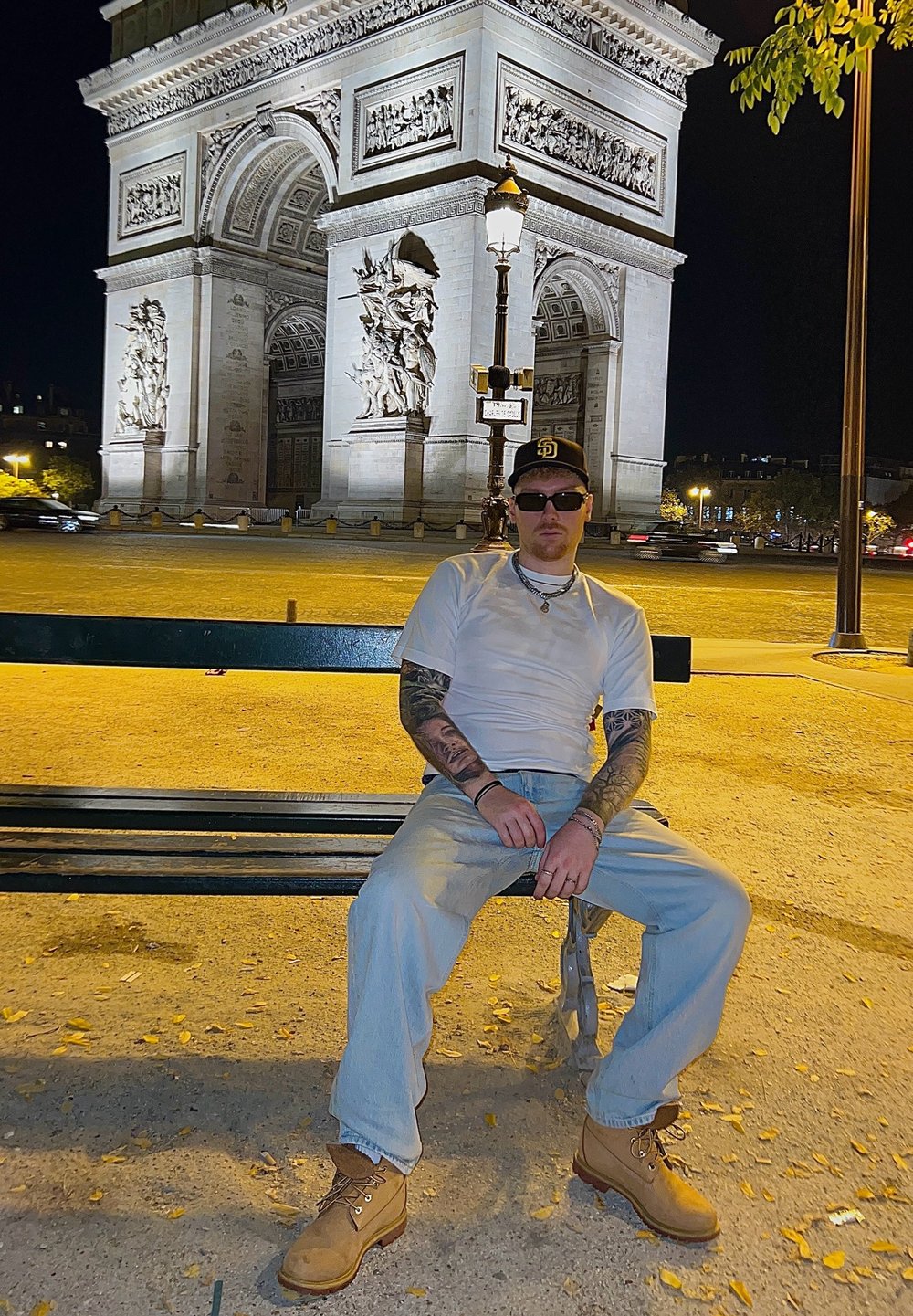 Persona seduta su una panchina verde scuro, con una maglietta bianca e jeans azzurri, stivaletti da lavoro marrone chiaro e occhiali da sole, davanti all'illuminato Arco di Trionfo.