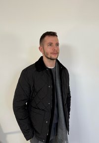 Homme portant une veste matelassée noire, un pull gris et une écharpe grise, debout contre un mur blanc uni, regardant légèrement vers le haut.