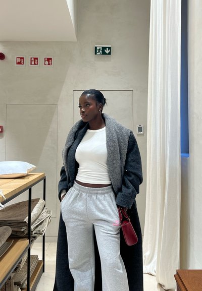 Mujer con abrigo gris sobre camiseta blanca y pantalones de chándal grises, de pie en el interior cerca de telas dobladas y cortinas claras.
