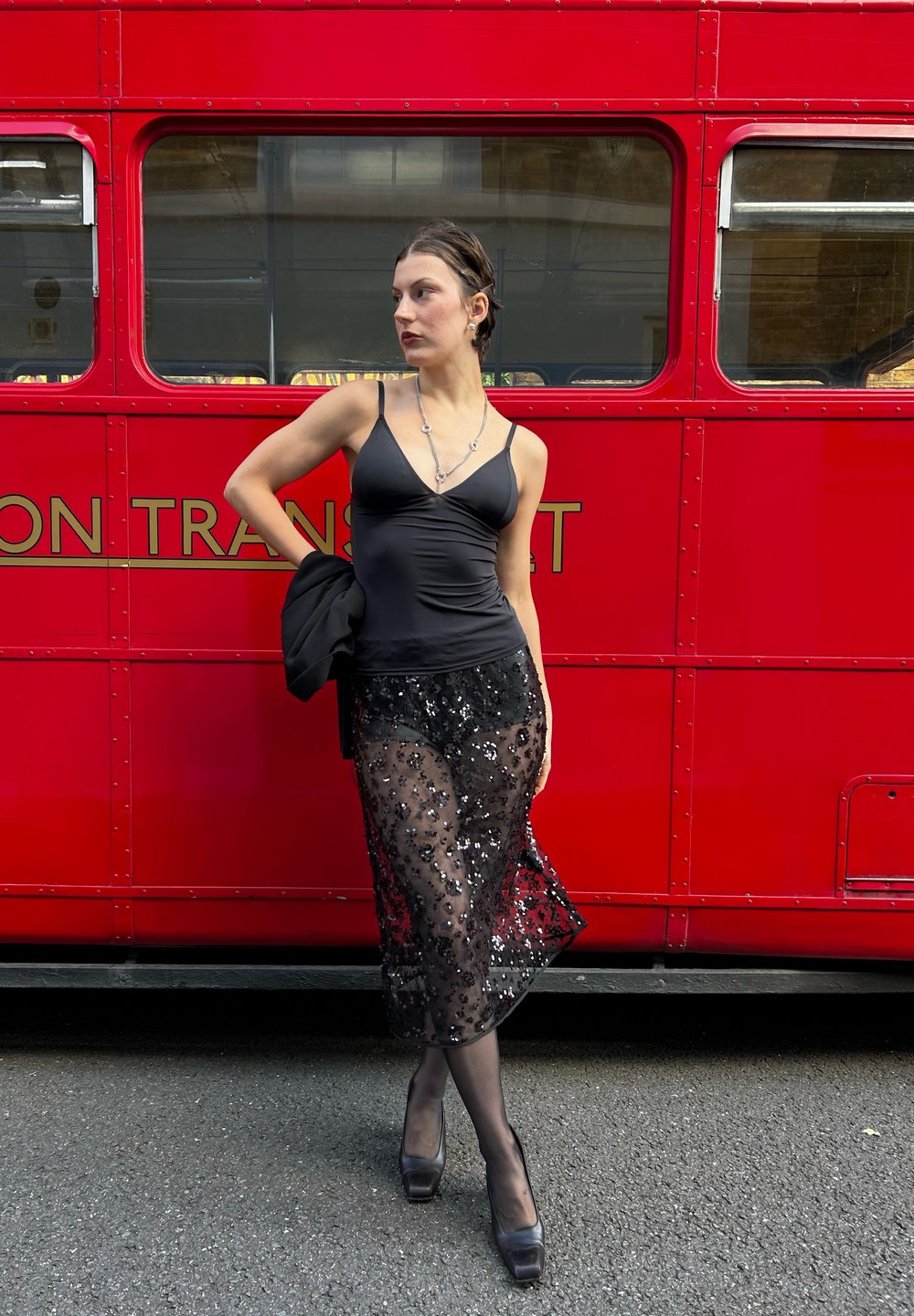 Černá saténová top bez rukávů doplněná průhlednou černou sukní posetou flitry. Oblečení doplňují černé lodičky na vysokém podpatku a černá bunda.