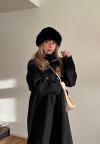 Kobieta o długich blond włosach, ubrana w czarny futrzany kapelusz i czarny płaszcz, trzyma beżową torebkę, stoi wewnątrz na tle jasnej ściany.