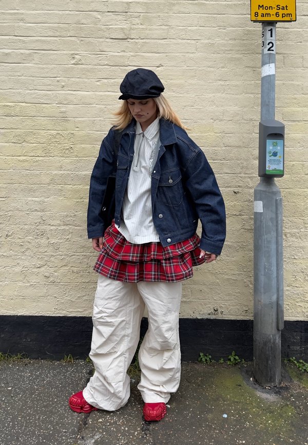 Young person wearing a black cap, dark jacket, white shirt, red plaid skirt over white pants, and red shoes standing near a street pole.