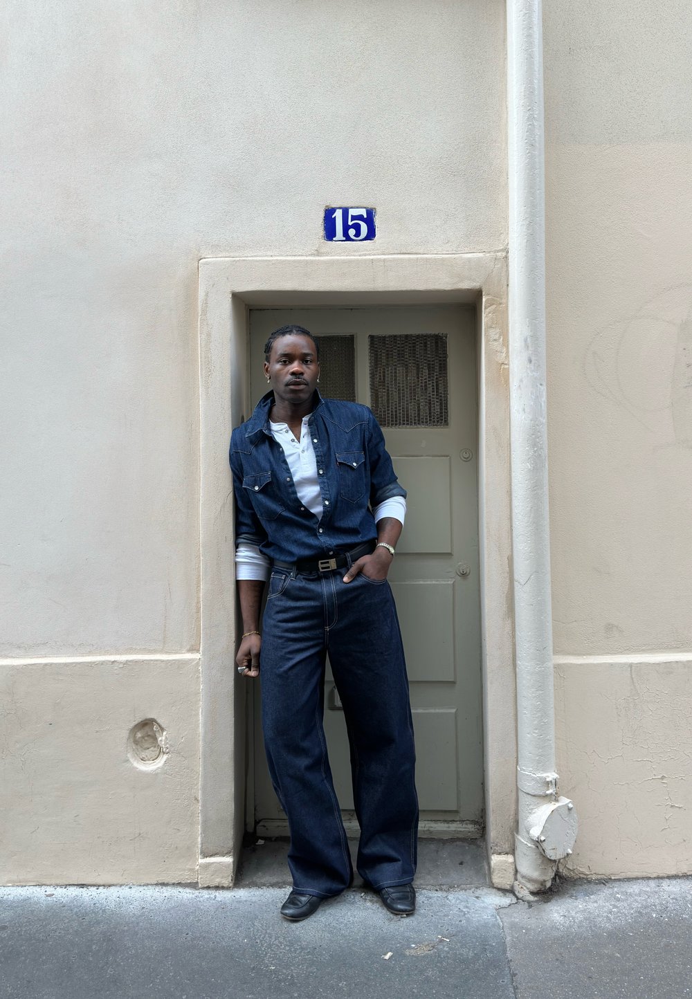 Personne portant un jean et une veste en denim foncé, se tenant dans une petite porte sous un panneau bleu avec le numéro 15 sur un mur beige.