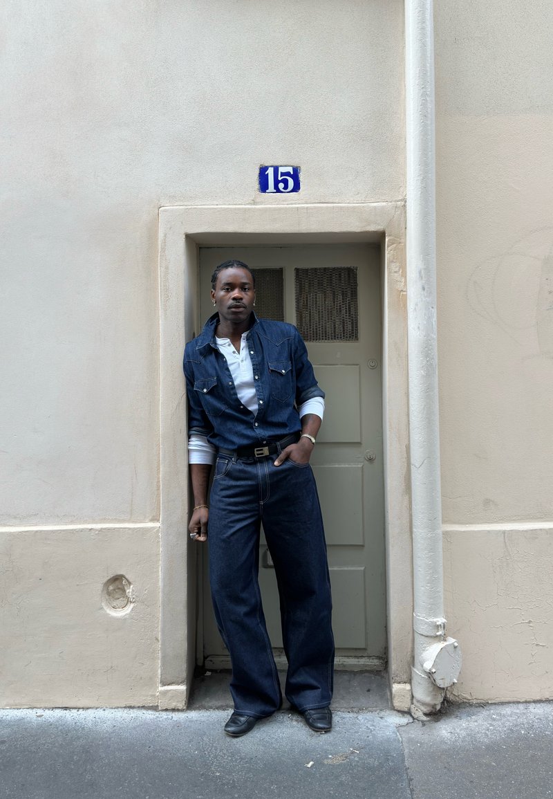 Personne portant un jean et une veste en denim foncé, se tenant dans une petite porte sous un panneau bleu avec le numéro 15 sur un mur beige.
