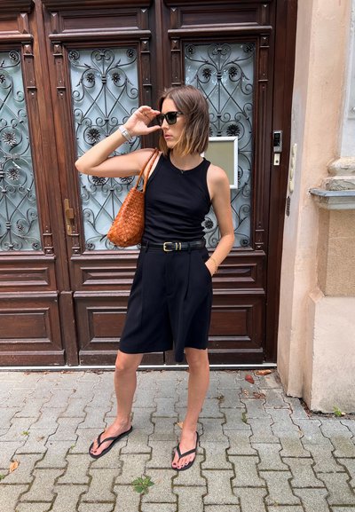 Top negro sin mangas, pantalones cortos pleat negros, bolso de mimbre naranja, gafas de sol oscuras y chanclas negras se encuentran frente a una puerta de madera con detalles ornamentales.