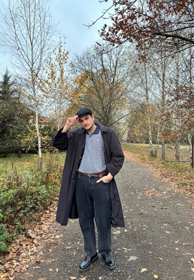 Abrigo negro, polo gris con patrón, jeans negros, cinturón marrón y zapatos negros. De pie en un camino entre árboles con hojas otoñales.