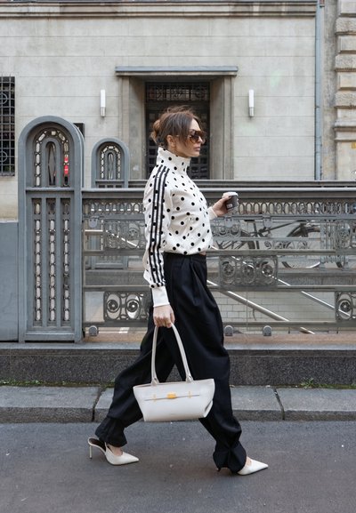 Mujer caminando por la calle de la ciudad con chaqueta de lunares, pantalones negros, tacones blancos, gafas de sol, llevando un bolso blanco y sosteniendo una taza de café.