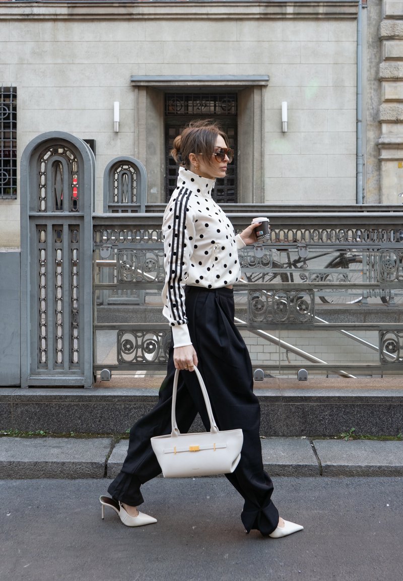 Žena hoda gradskom ulicom noseći jaknu s točkicama, crne hlače, bijele štikle, sunčane naočale, noseći bijelu torbu i držeći šalicu kave.