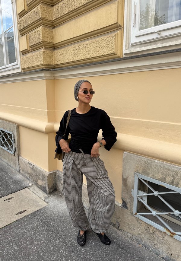 Mujer que lleva un top negro, pantalones anchos a cuadros, gafas de sol y pañuelo en la cabeza, de pie en una acera junto a una pared texturizada amarilla de un edificio.