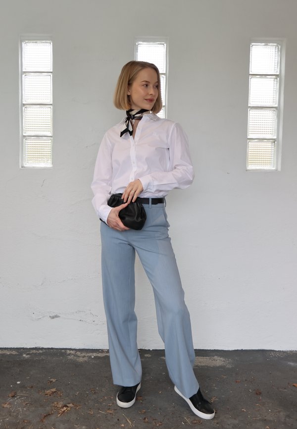 Vrouw draagt wit overhemd, lichtblauwe broek, zwarte schoenen en een halsdoek, staat met een kleine zwarte handtas tegen een witte muur met glasblokramen.