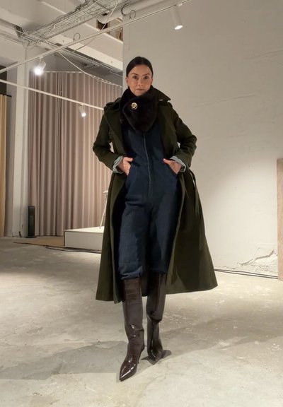 Mujer con un abrigo verde oscuro, mono azul marino, botas marrones hasta la rodilla y una gran bufanda negra con un broche dorado, dentro de una habitación minimalista.