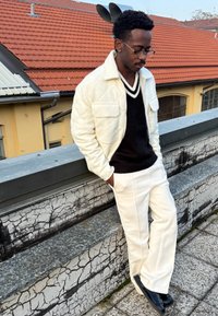 Giacca bianca trapuntata, maglione nero a scollo a V, pantaloni bianchi a gamba larga e scarpe nere. In piedi di fronte a uno sfondo testurizzato grigio e pietra.