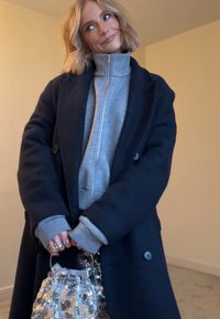 Manteau en laine bleu marine sur un sweat à capuche gris zippé, tenant un sac à main argenté à sequins avec un cordon de serrage. Textures douces et matériaux mélangés visibles.