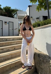 Fekete bikini felső, fehér vászonnadrág és fekete flip-flop szandál. Narancssárga ital a kezében, kövekből készült lépcsőkön áll egy napos szabadtéri környezetben.