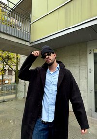 Mees, kellel on peas must beanie, prillid, must mantel, helesinine särk ja teksad, seisab õues betoonhoone lähedal kerges vihmas.