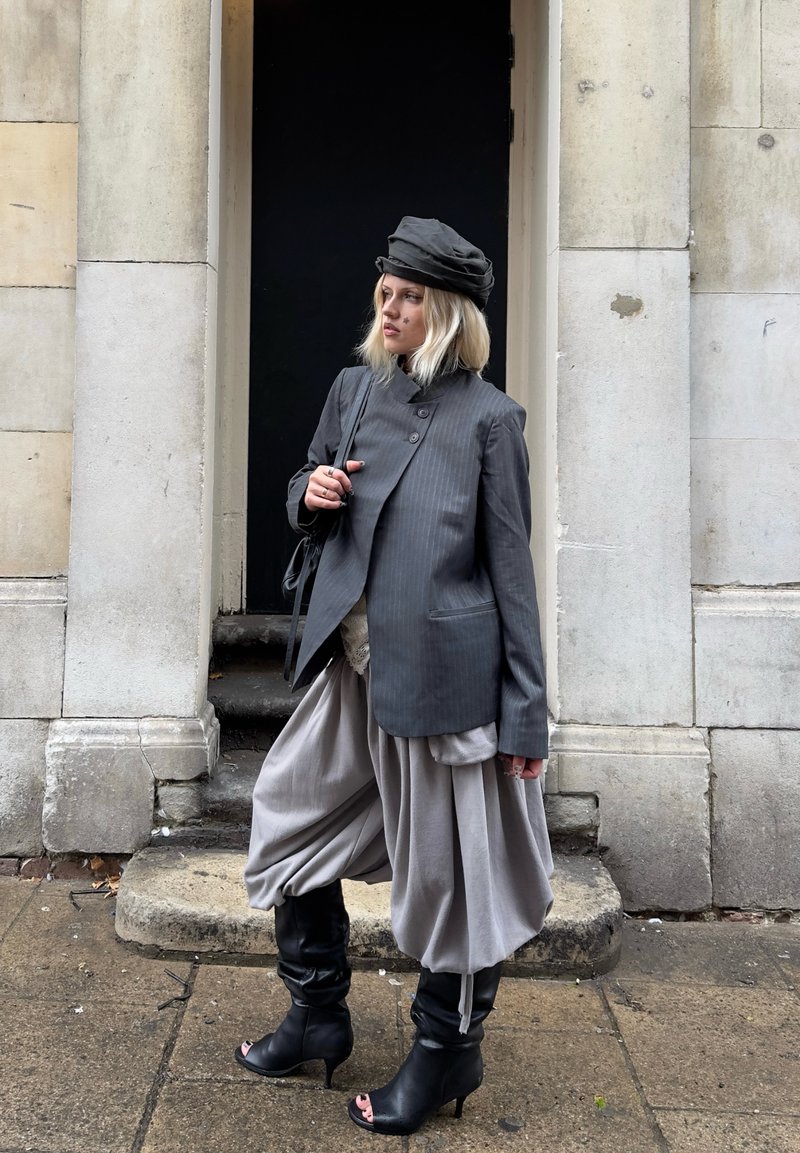Femme blonde debout sur des marches en pierre, portant un blazer gris à fines rayures, un pantalon gris volumineux, des bottines noires à bout ouvert et un chapeau gris assorti.