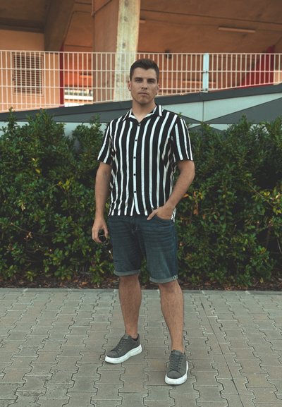 Camisa de manga corta a rayas verticales en blanco y negro, pantalones cortos de mezclilla, zapatillas grises, de pie sobre un área pavimentada con vegetación de fondo.