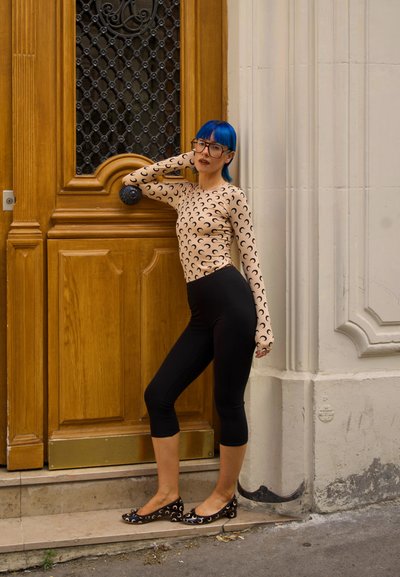 Top beige de manga larga con patrón de media luna negra, leggings capri negros y zapatos planos negros con detalles de media luna, posando junto a una puerta de madera.