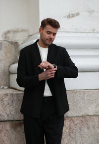 Juodas švarkas ant baltos marškinėlių, matomas sidabrinis žiedas ir juodas laikrodis ant riešo. Fone yra tekstūruotas akmuo.