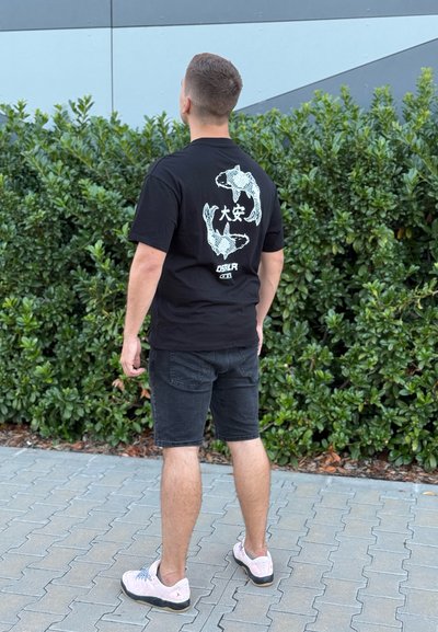Camiseta negra con un gráfico de un pez koi blanco y texto en la parte posterior. Lleva puesta un pantalón corto negro y zapatillas de deporte rosa claro. Hombre visto desde detrás.
