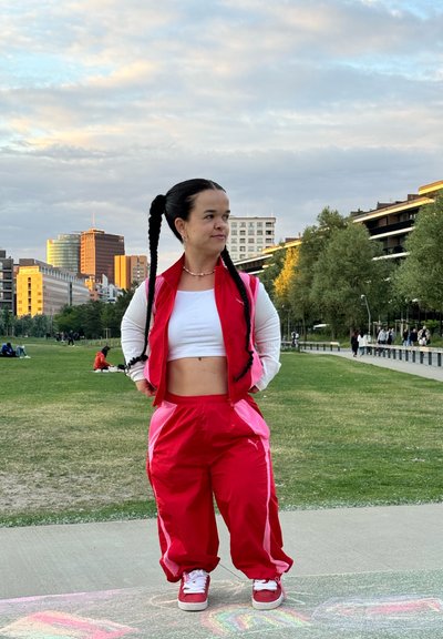 Chándal deportivo rojo con acentos blancos, top corto y zapatillas a juego. Cabello largo y trenzado, de pie en una zona de césped con edificios al fondo.