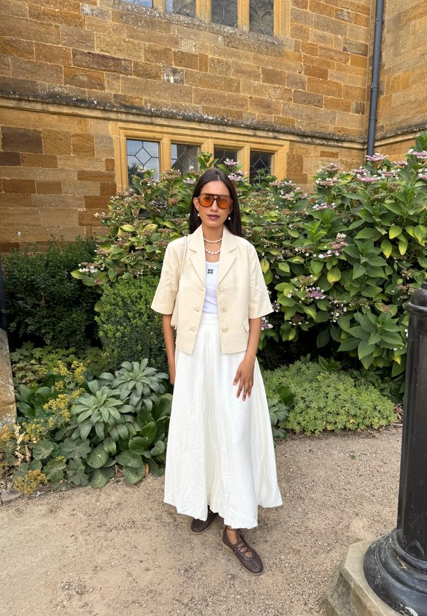Donna che indossa un blazer crema, top bianco, gonna lunga bianca e scarpe marroni, in piedi di fronte a un edificio di mattoni con cespugli verdi e fiori.