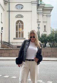 Black leather jacket with fur cuffs, white shirt, beige trousers, and sunglasses; standing against a light-coloured building with large windows.