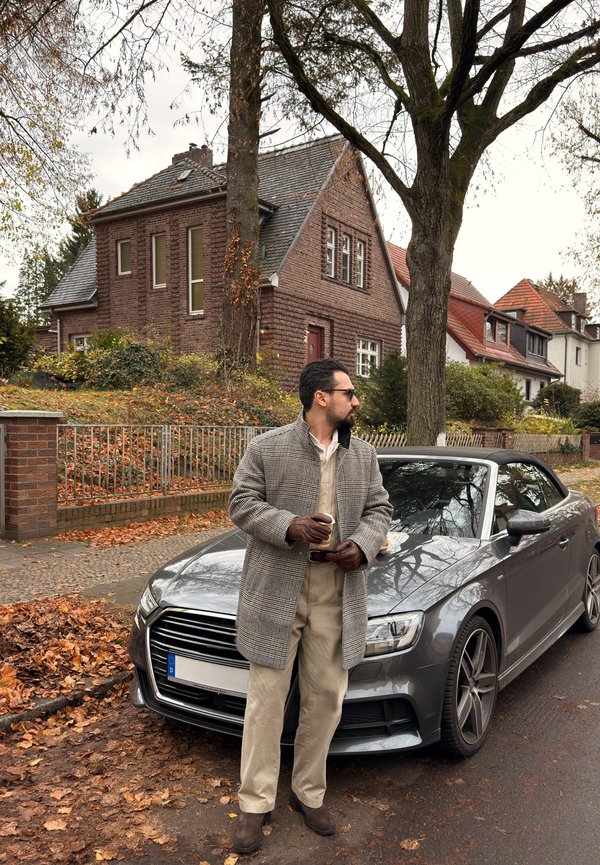Coche descapotable gris estacionado en una calle con hojas de otoño. El sujeto lleva un abrigo de pata de gallo, pantalones beige y guantes marrones, sosteniendo una taza.