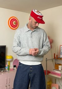 Sivi pleteni pulover z ovratnikom, oblečen čez belo srajco, kombiniran z razširjenimi modrimi denim hlačami. Nošena je rdeča kapa s krožnim grafikonom.