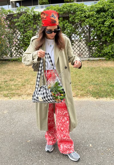 Mujer con gabardina beige, pantalones rojos con estampado, pañuelo rojo con dibujos de perros, gafas de sol, sosteniendo un bolso a cuadros con flores al aire libre.