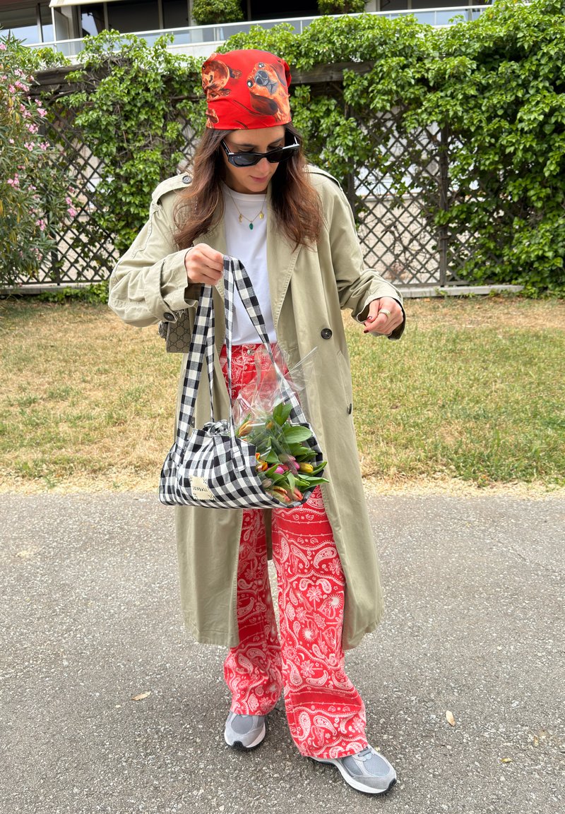 Femme portant un trench-coat beige, pantalon rouge à motifs, foulard rouge avec imprimés de chiens, lunettes de soleil, tenant un sac à carreaux avec des fleurs en extérieur.