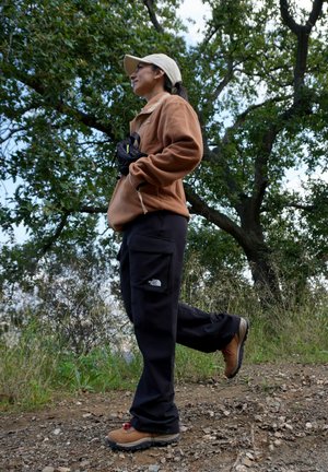 Persona con una chaqueta de forro polar color beige, pantalones cargo negros con bolsillos laterales y botas de senderismo marrones con cordones rojos, caminando por un sendero de tierra.